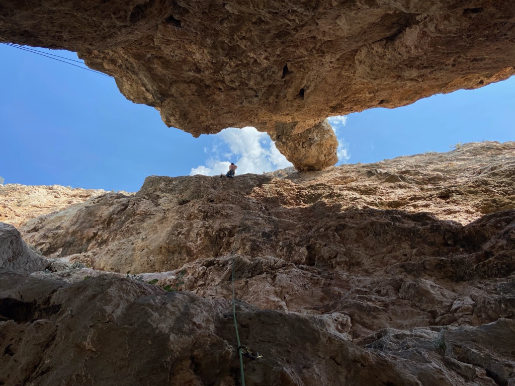 Climber in-between the cliff and the pinnacle. There is blue sky and clouds appearing between the gap in the roof.