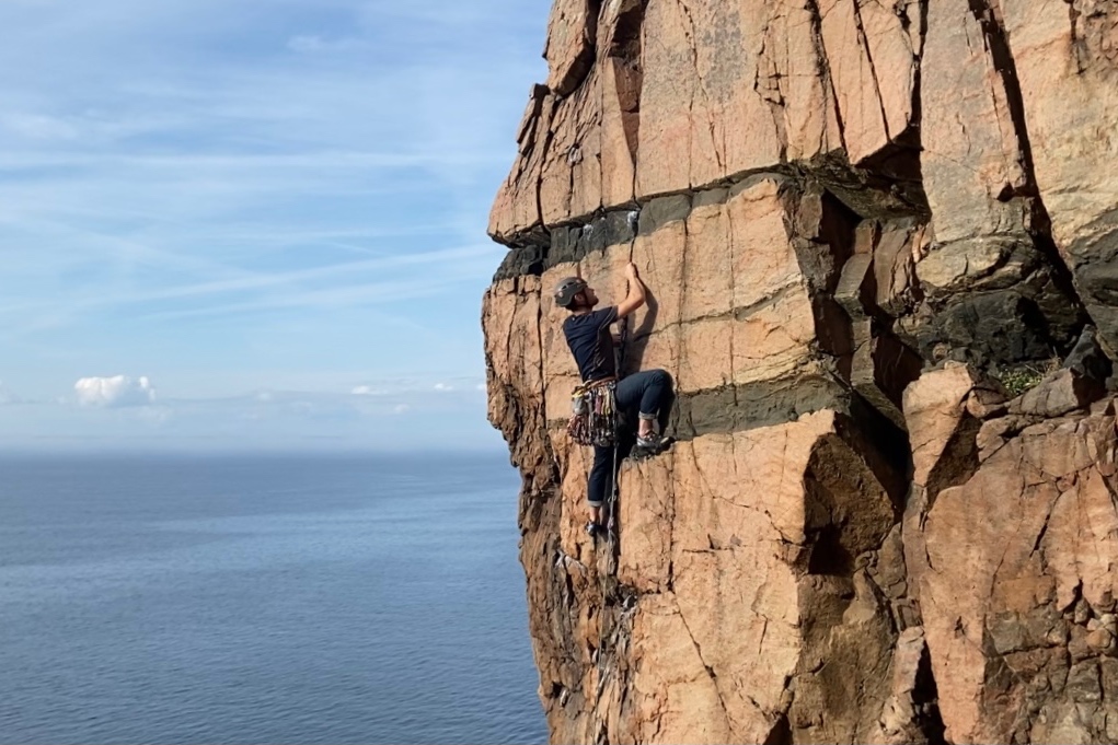 Climber on pink granite which has horizontal grey/black striations. In the background is the sea
