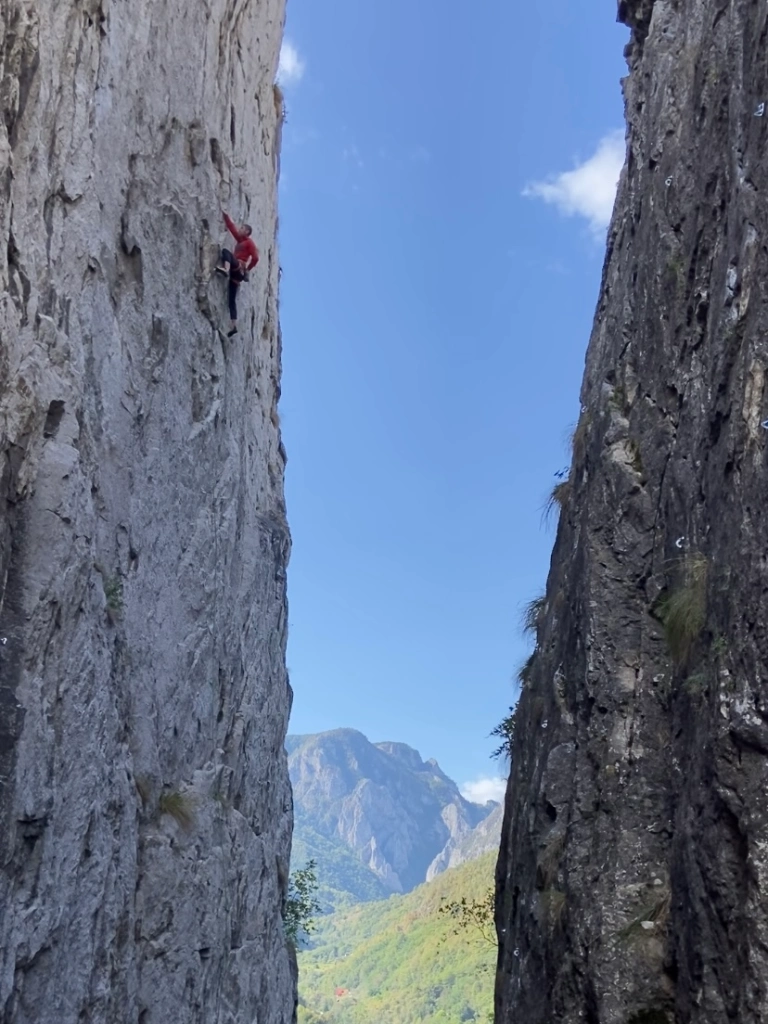 Climber chalking up on a tall vertical cliff on the left hand side of a narrow canyon. Between the two walls the view looks over a forested valley and another rocky limestone massif 