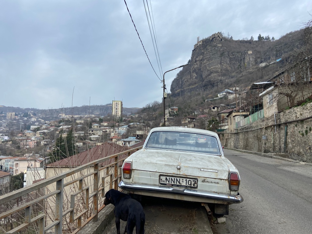 Old soviet era Volga saloon car parked up on a street with power lines overhead with black cliff set upon the hillside. 