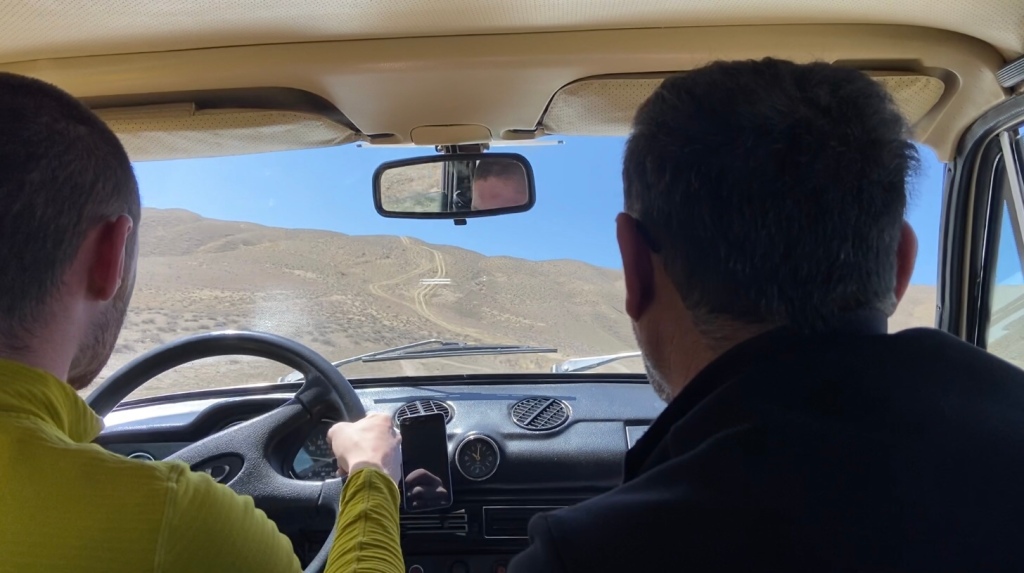 Looking out from the rear passenger seats of an old soviet era Lada with two people in the front seats driving off road up a steep hill 