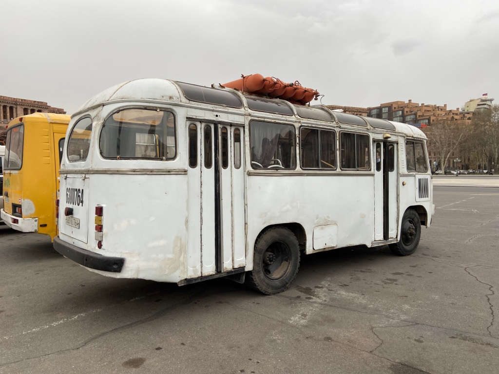 Old white bus with rounded windows and six large orange LPG tanks strapped to the roof. The old folding doors are half open 